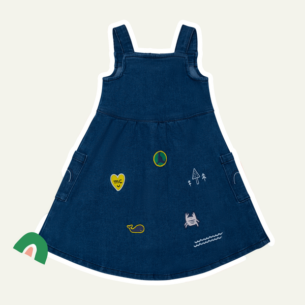 A navy blue denim baby dress with decorative patches on the sleeves and hem, including a heart and other shapes, displayed against a white background with pink accents.