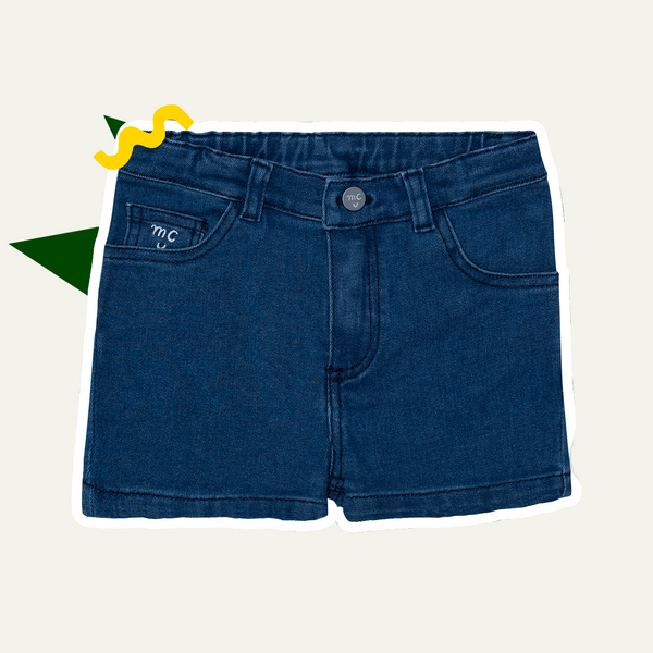Navy blue denim girl shorts with a graphic label on the left leg and a decorative yellow tag on the right leg, displayed against a white background with a green and yellow explosion graphic behind it.